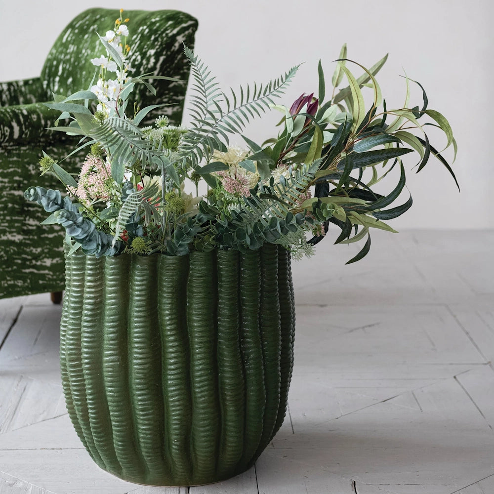Green fluted stoneware planter filled with various faux greenery and flowers on a light wood floor.