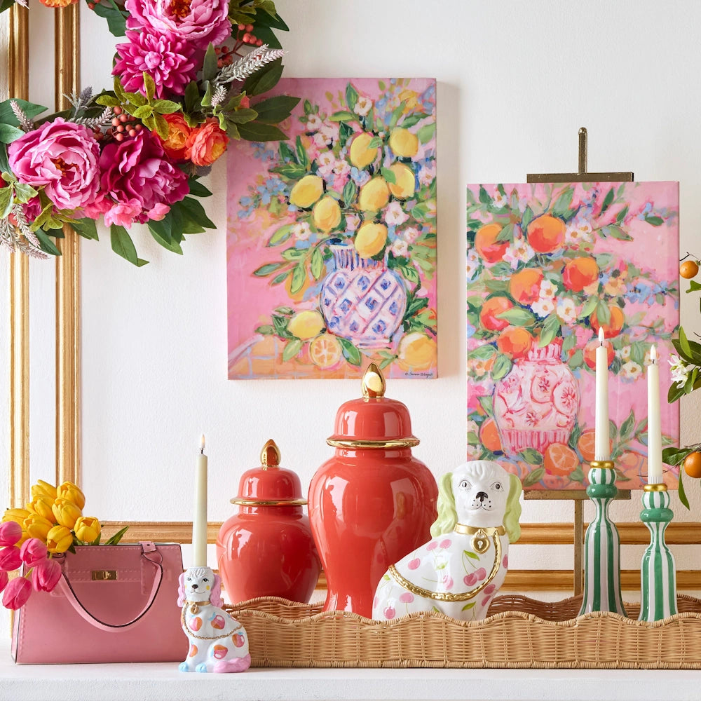 Vibrant floral wreath, two colorful floral paintings on pink backgrounds, orange jars, ceramic dogs, pink handbag, and candles on a wicker tray.