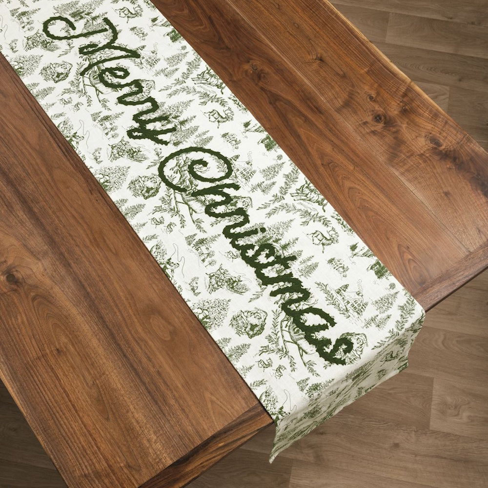 Green toile table runner with "Merry Christmas" script laid across a wooden dining table.