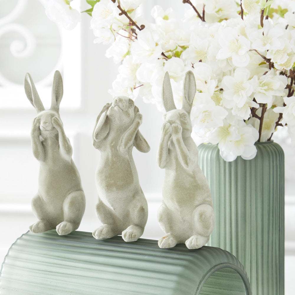 Set of three green velvet bunny figurines depicting hear, see, and speak no evil poses next to white flowers in a vase.