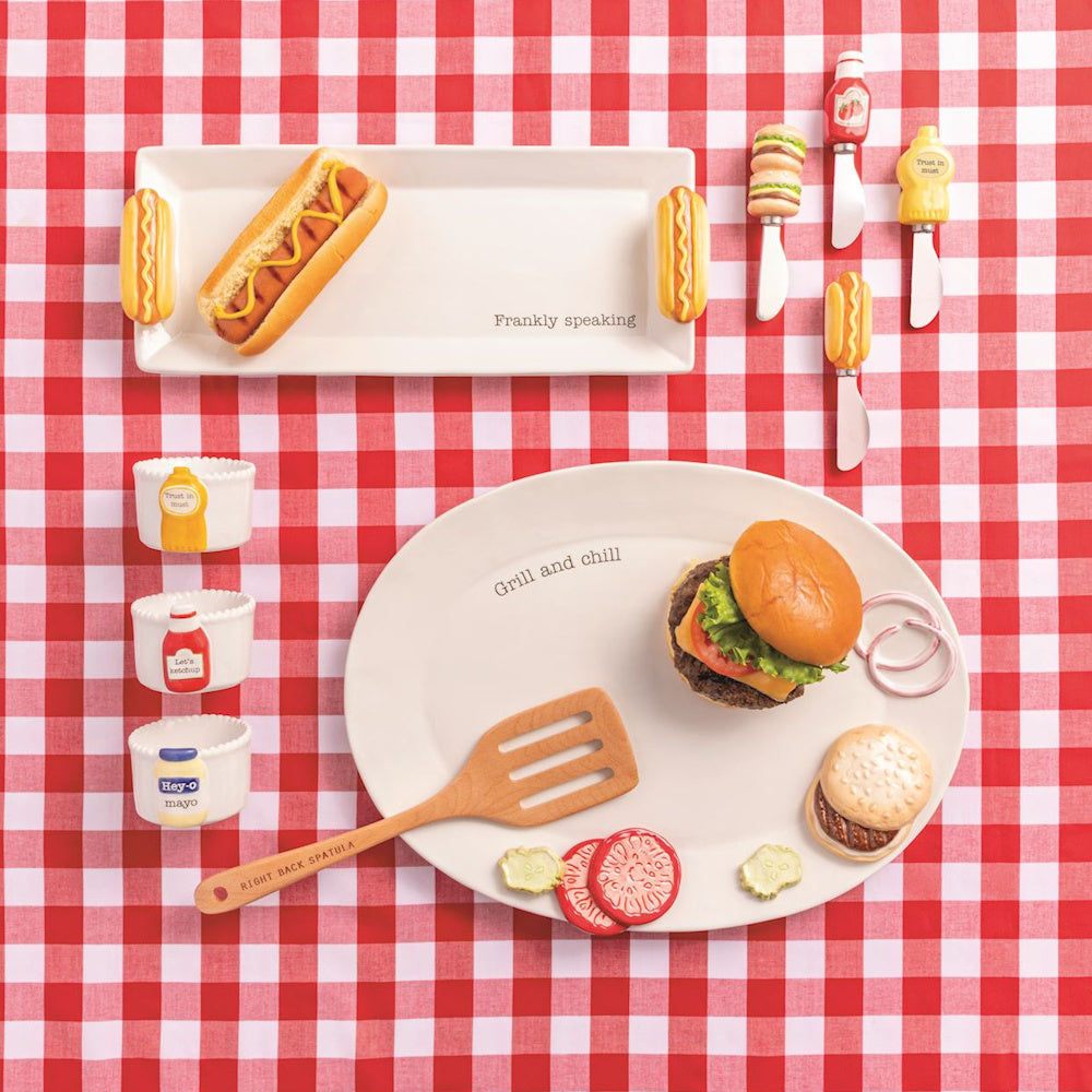 BBQ-themed ceramic serving set with hot dog platter, burger plate, condiment cups, and spreaders on red checkered fabric.