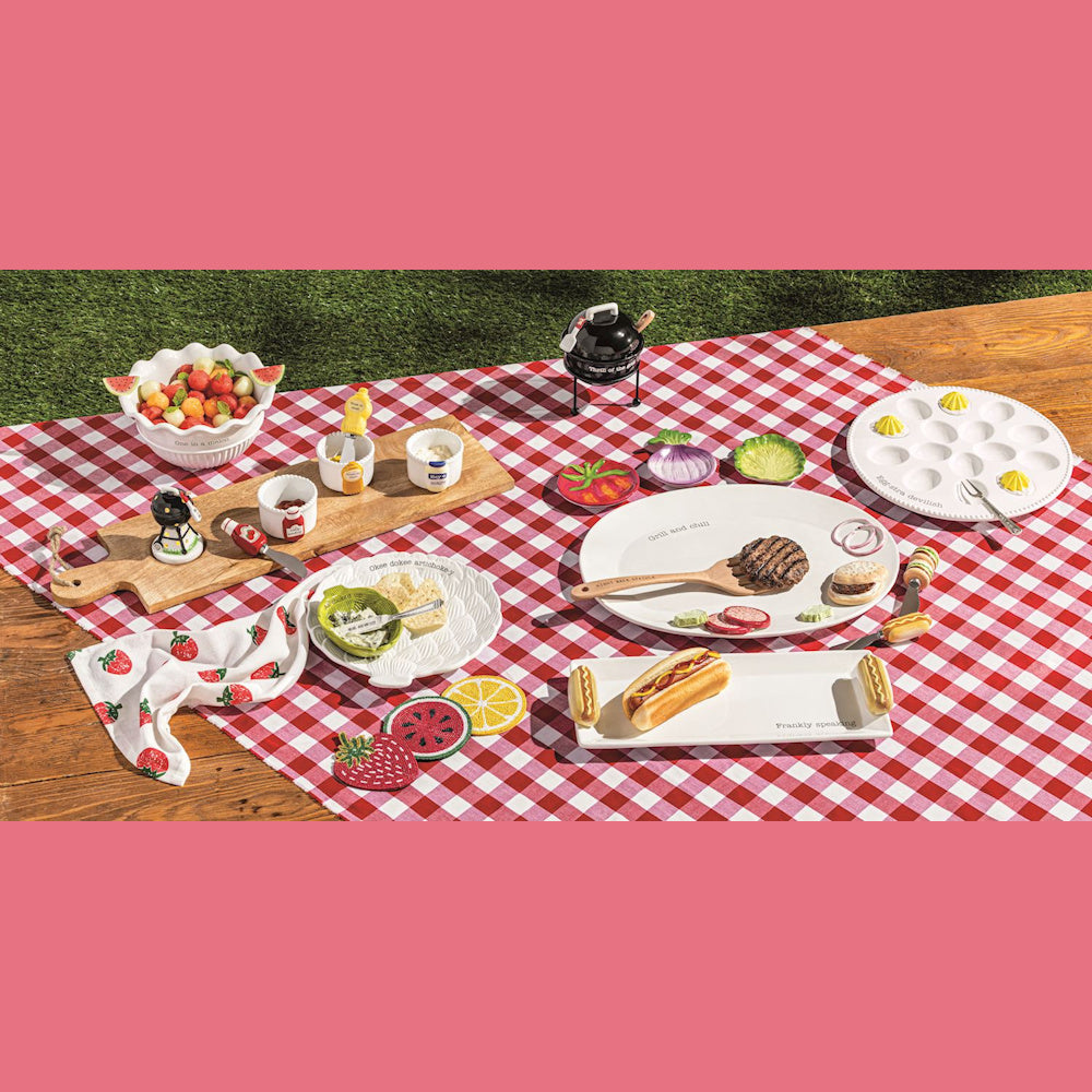 Summer picnic spread with BBQ-themed serving platters, condiment sets, and fruit bowls on a red gingham tablecloth.