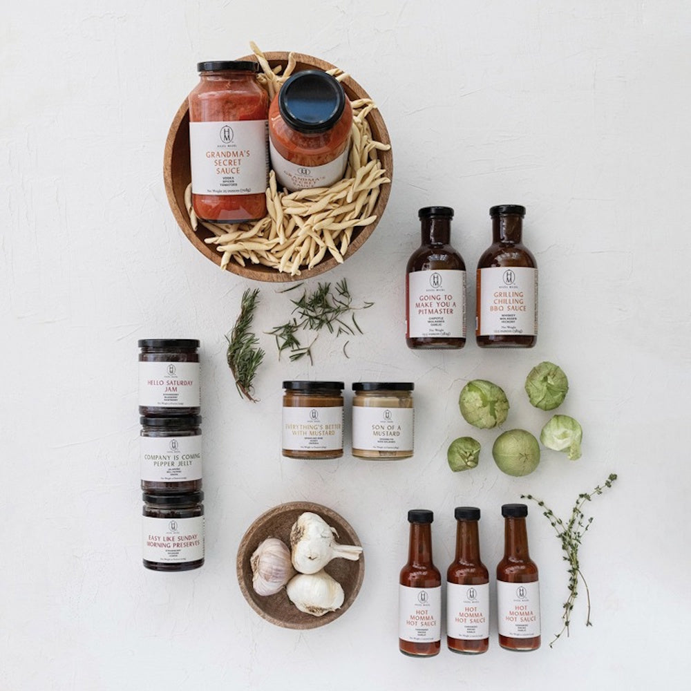 An assortment of gourmet sauces, jams, and mustards arranged with pasta, garlic, and herbs on a white surface.