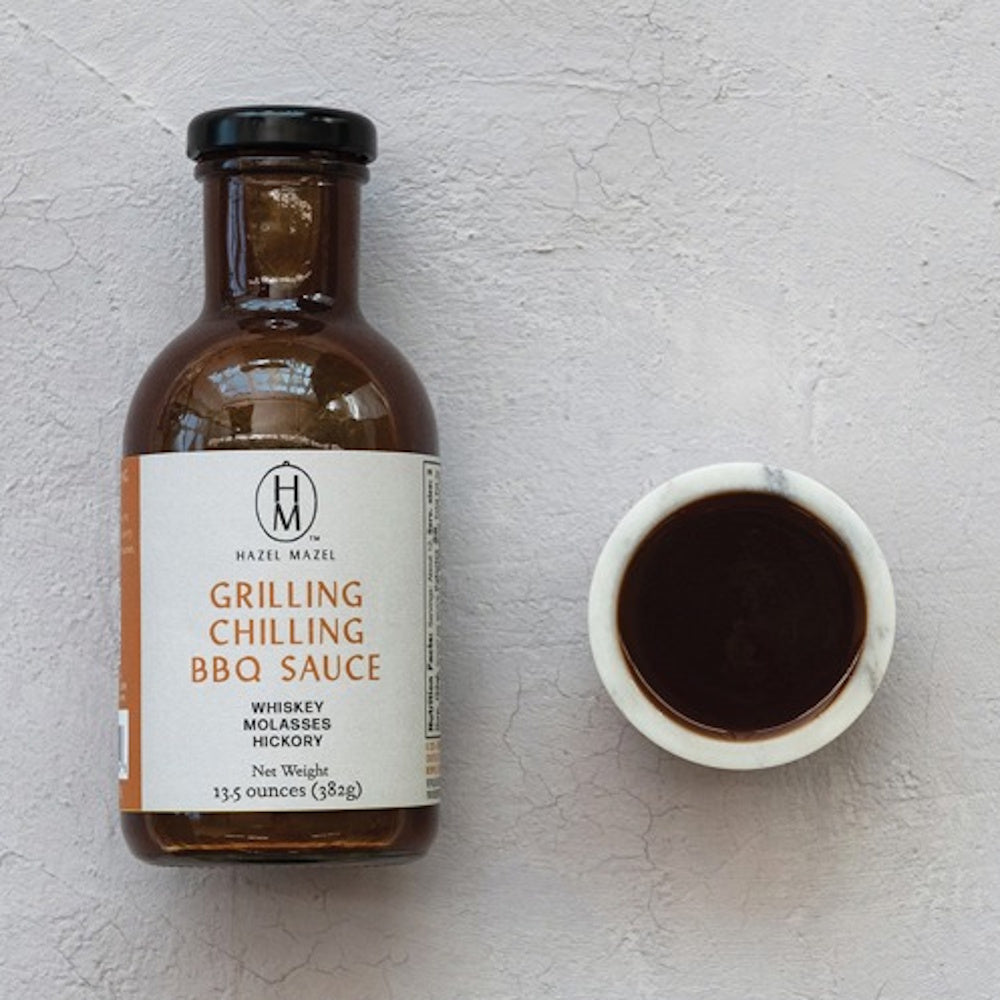 A bottle of Hazel Mazel Grilling Chilling BBQ Sauce next to a small marble bowl filled with the dark sauce.