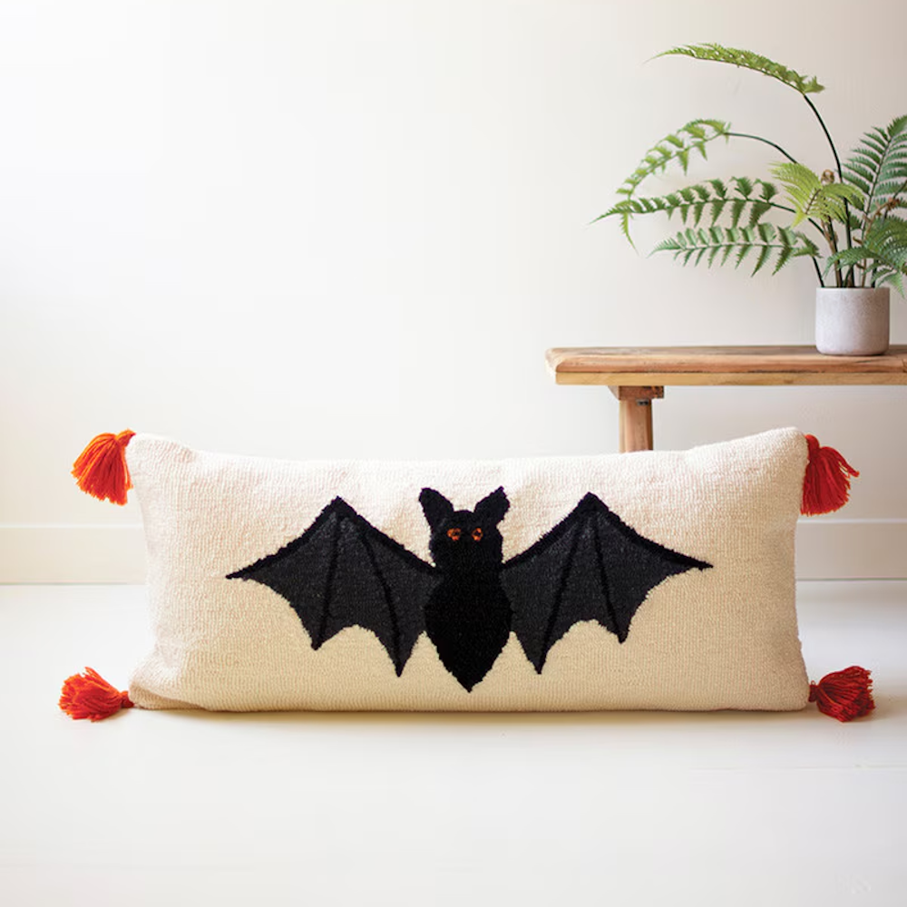 Cream hand-hooked lumbar pillow with a black Halloween bat and orange tassels on the corners.