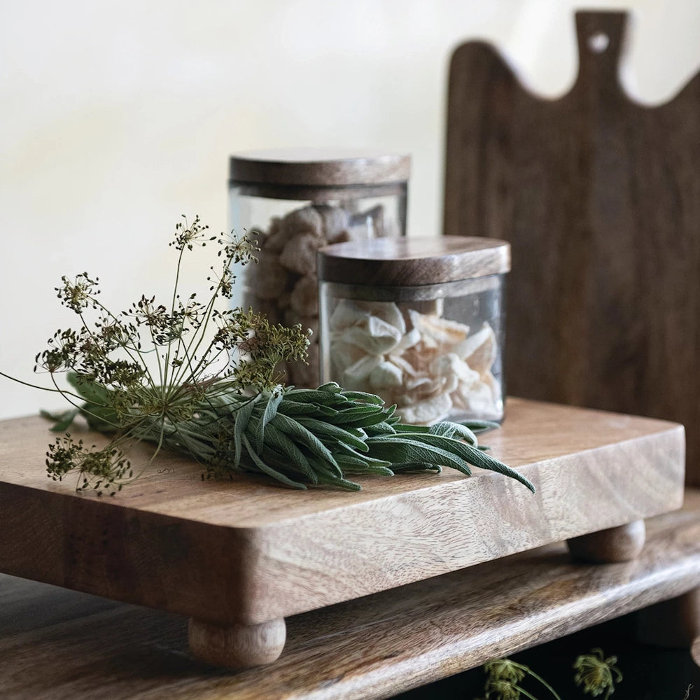 Hand-blown glass jars with mango wood lids on a wooden pedestal with fresh sage and dried herbs for kitchen storage.