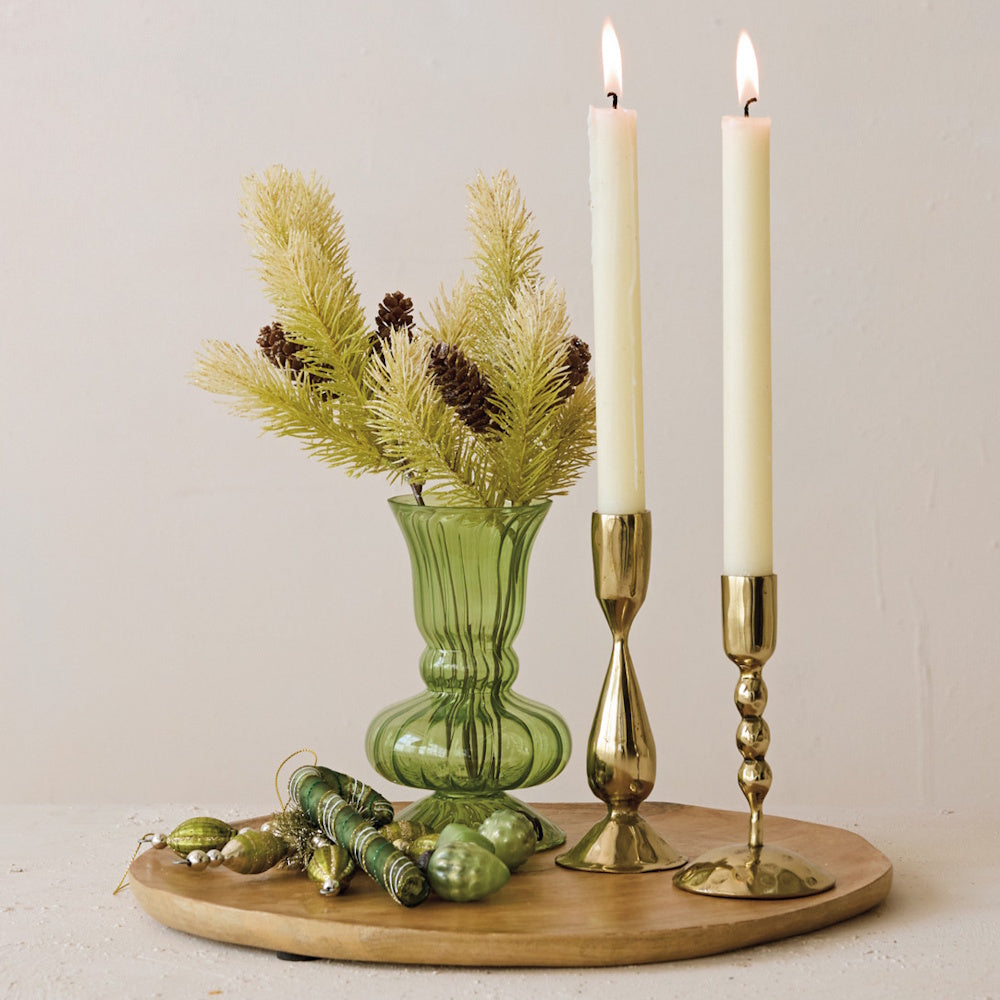 Two brass taper candle holders with lit white candles next to a green vase and ornaments on a wooden tray.