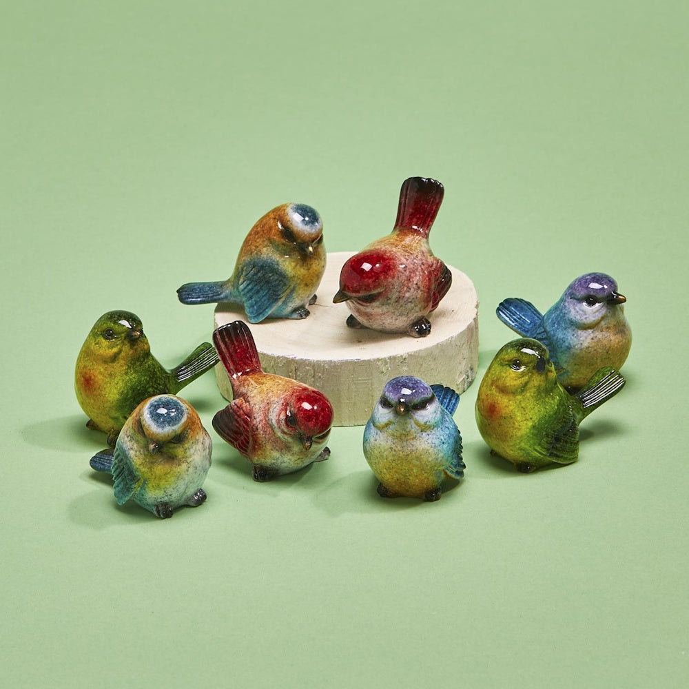 A collection of colorful hand-painted ceramic bird figurines arranged around a wood slice on a sage green background.