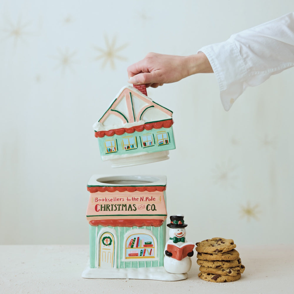 Hand-painted ceramic bookstore-shaped Christmas cookie jar with its lid lifted off by a hand, beside a stack of cookies.