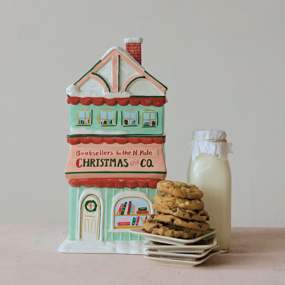 Hand-painted ceramic bookstore-shaped Christmas cookie jar displayed with a stack of cookies and a bottle of milk.