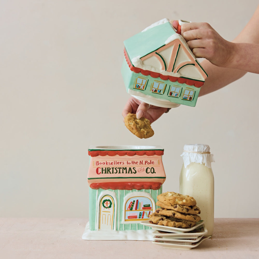Hand-painted ceramic bookstore-shaped Christmas cookie jar being opened to reveal a cookie inside.