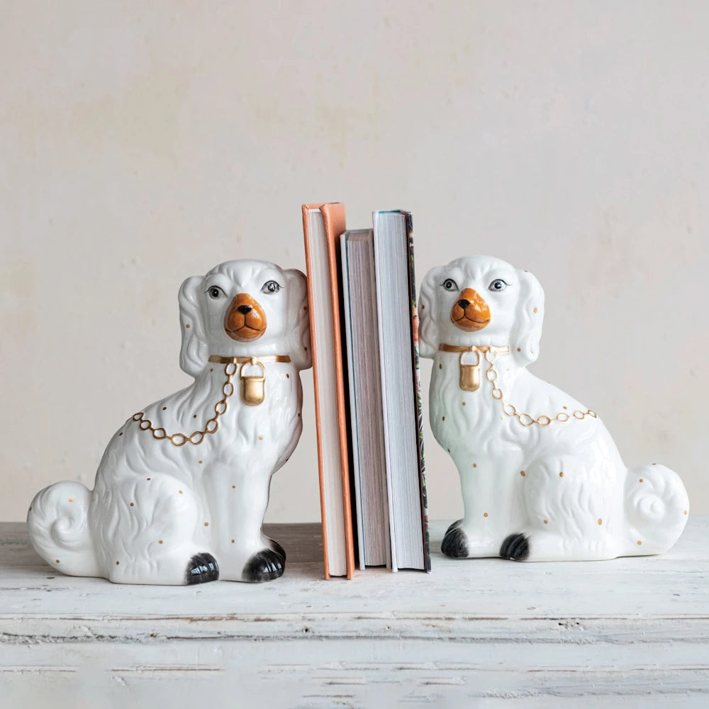 White hand-painted Staffordshire dog bookends with gold accents holding books on a rustic white wooden shelf.