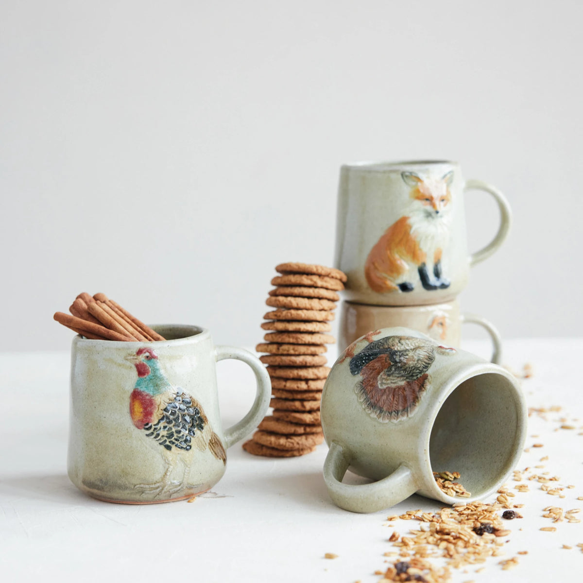 Animal-themed ceramic mugs arranged with cinnamon sticks, cookies, and spilled granola on a white surface.