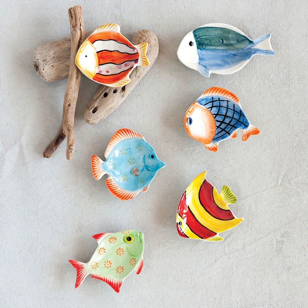 Six hand-painted, fish-shaped ceramic dishes in vibrant colors arranged on a light textured surface with driftwood.