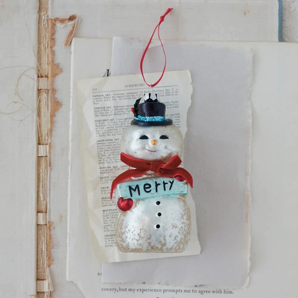 Hand-painted mercury glass snowman ornament wearing a top hat and holding a "Merry" sign, displayed on vintage book pages.