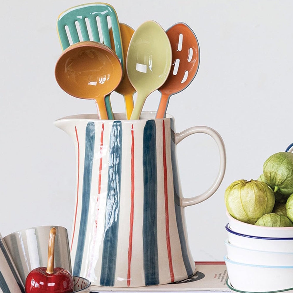 A hand-painted striped ceramic pitcher holds colorful kitchen utensils next to a stack of bowls with tomatillos.