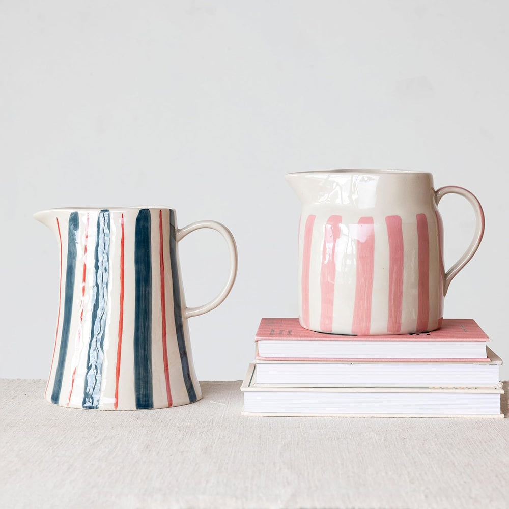 Two ceramic pitchers with hand-painted stripes, one blue and red, the other pink on a stack of books.