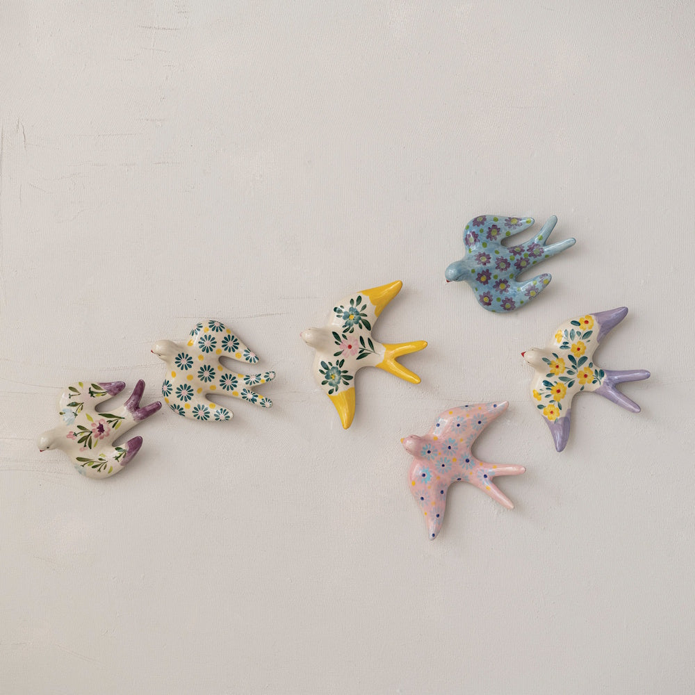 Six colorful hand-painted stoneware birds with floral patterns arranged as if flying across a white wall.