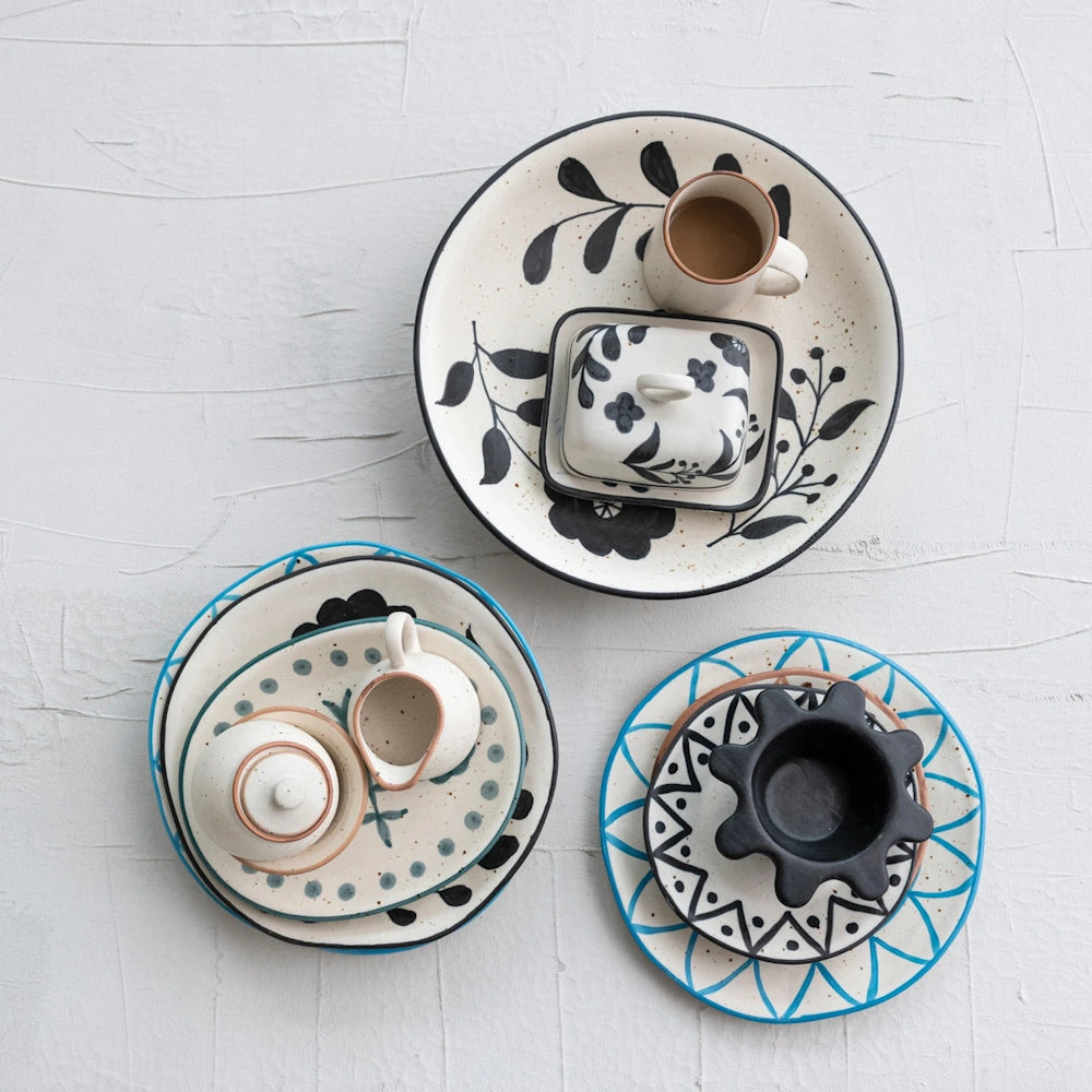 Flat lay view of a hand-painted floral stoneware butter dish styled alongside matching tableware on white wood.