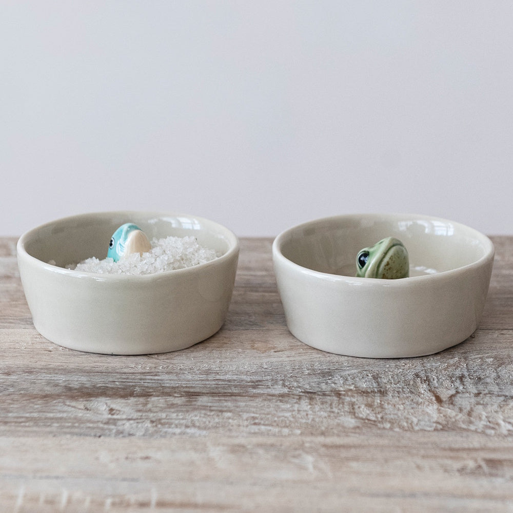 Two small stoneware dishes on a wooden table, one filled with salt and featuring a fish, the other a frog.