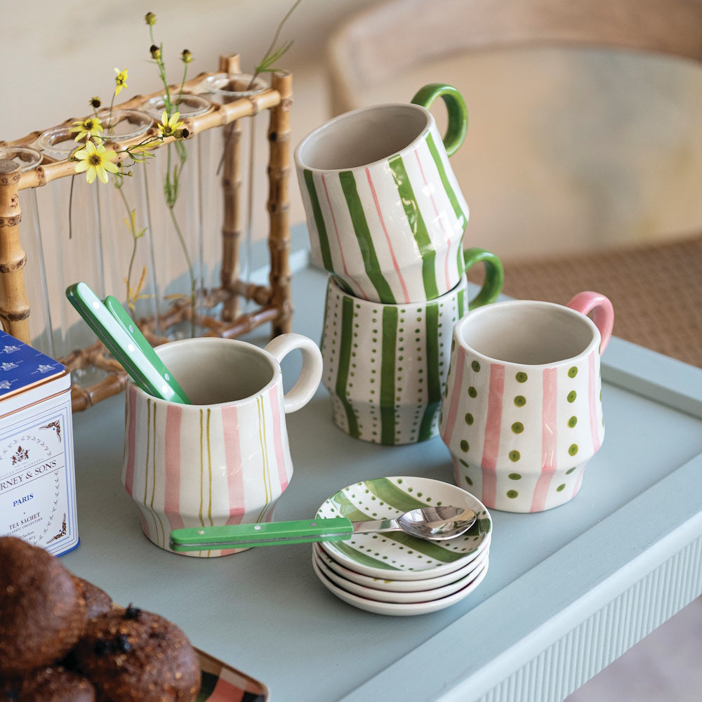Hand-painted ceramic mugs, mini saucers, and tea utensils arranged on a light blue table set for tea.