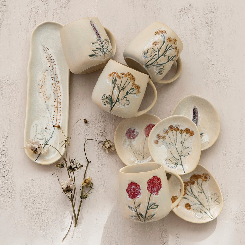 Lifestyle view of hand-painted stoneware mugs, plates, and a long tray featuring floral designs.