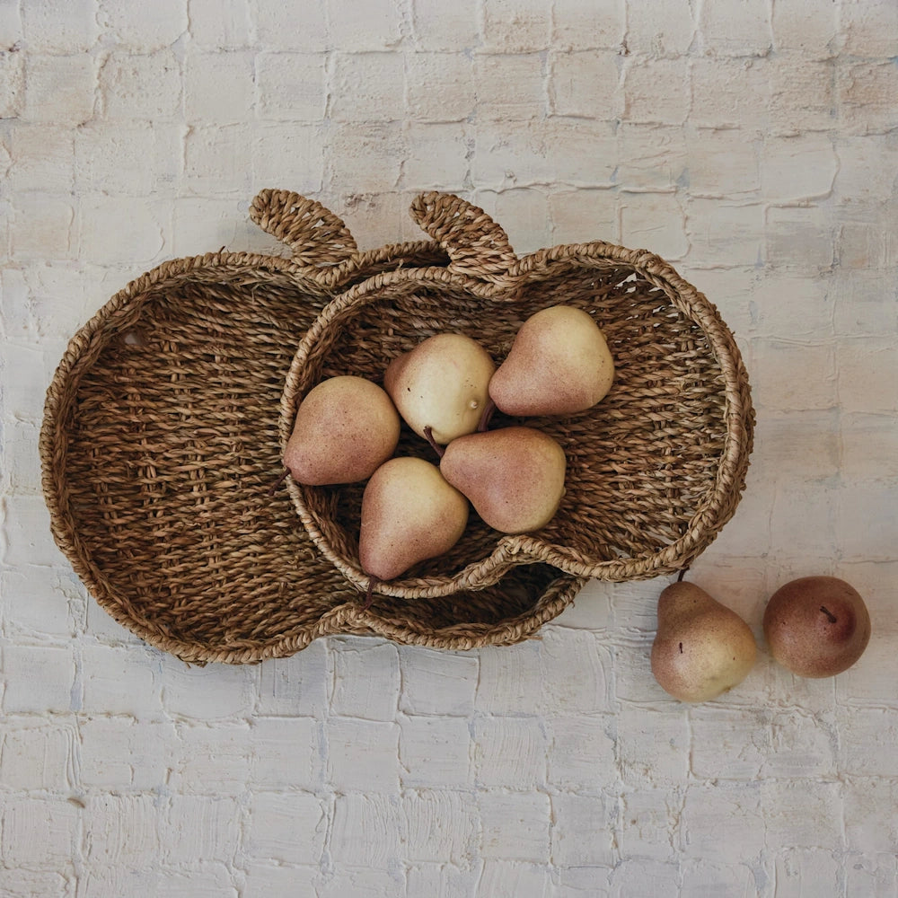 Top-down view of hand-woven pumpkin-shaped seagrass trays filled with fresh pears on a white textured background.
