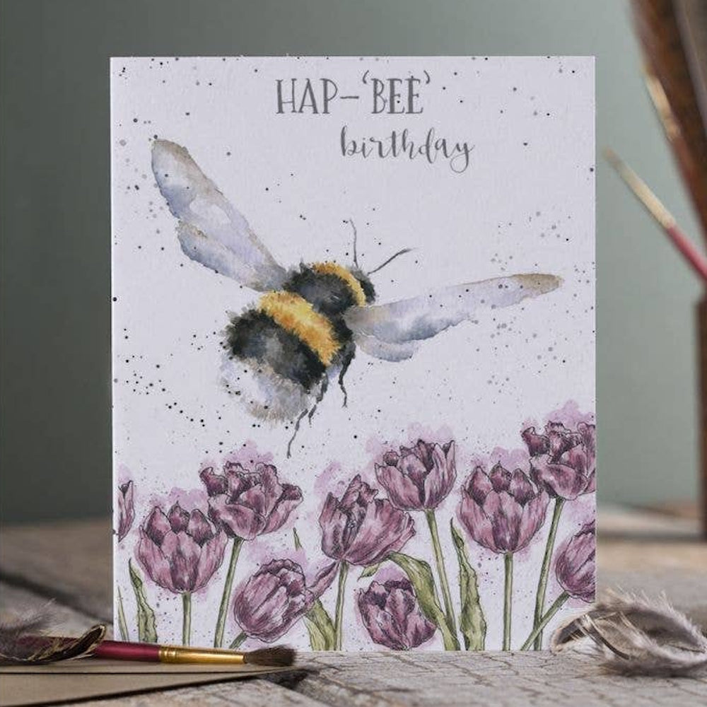 Watercolor bumblebee flying over purple tulips on a "Hap-Bee Birthday" card with paintbrushes in the foreground.