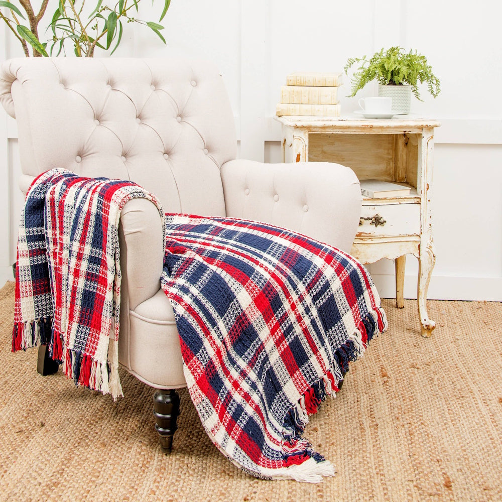 A red, white, and blue plaid throw blanket with fringed edges draped over a tufted cream-colored armchair.