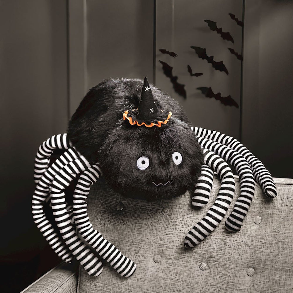 Plush black spider toy with striped legs and a tiny witch hat resting on a chair near bat decor.
