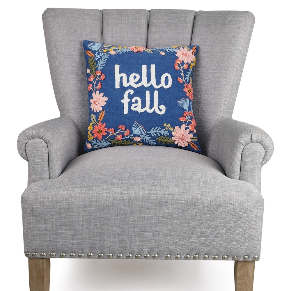 A blue embroidered pillow reading "hello fall" with floral borders sits on a gray armchair.