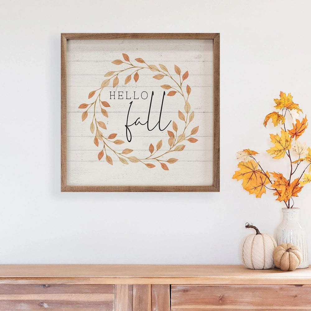 Framed "HELLO fall" sign hanging on a white wall above a wooden table with fall decor.