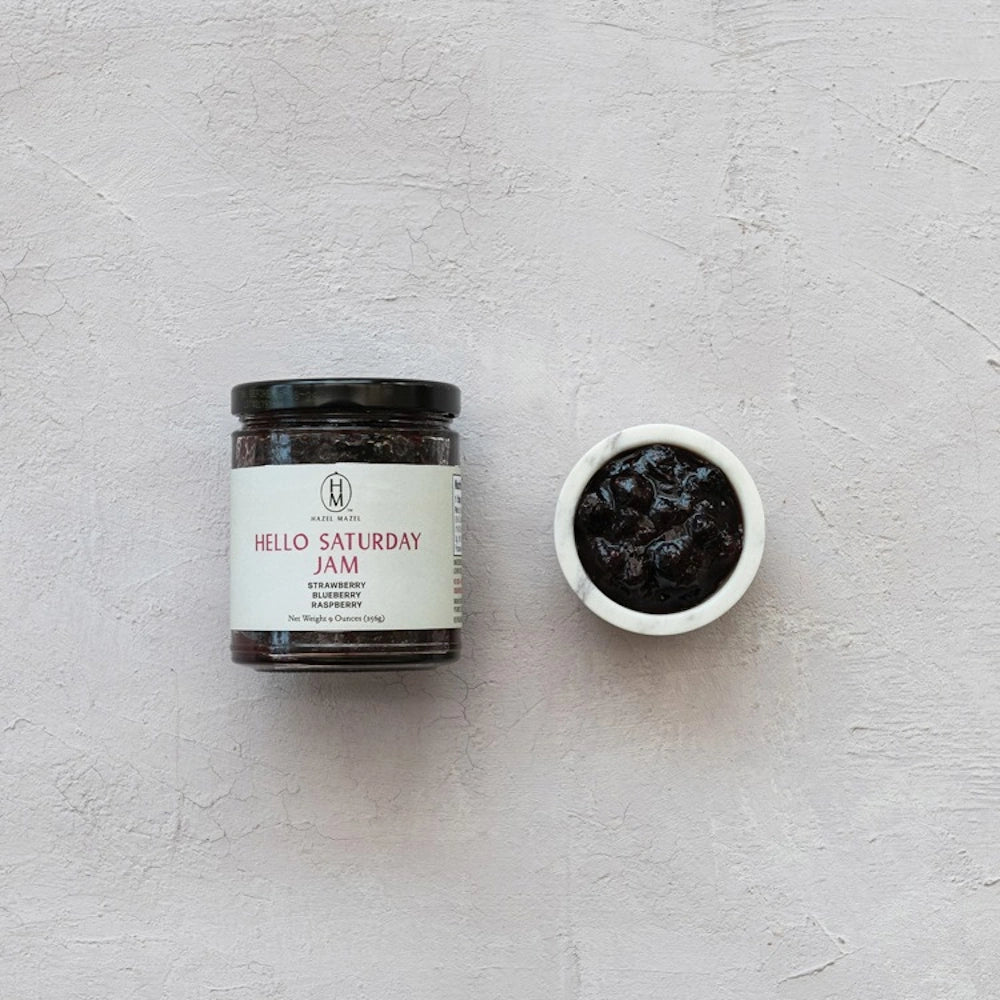 A jar of Hazel Mazel Hello Saturday Jam and a small marble bowl of mixed berry jam on a light grey background.