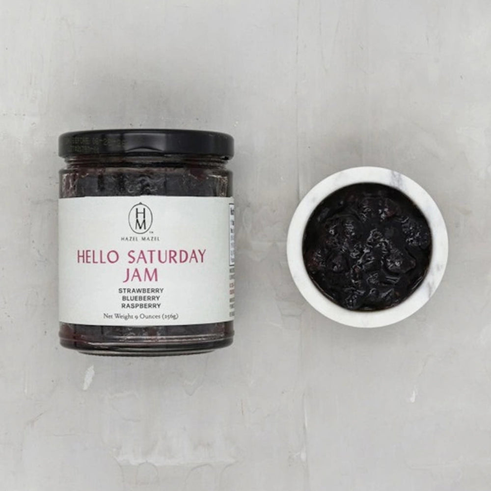 A jar of Hazel Mazel Hello Saturday Jam containing strawberry, blueberry, and raspberry next to a small bowl of jam.