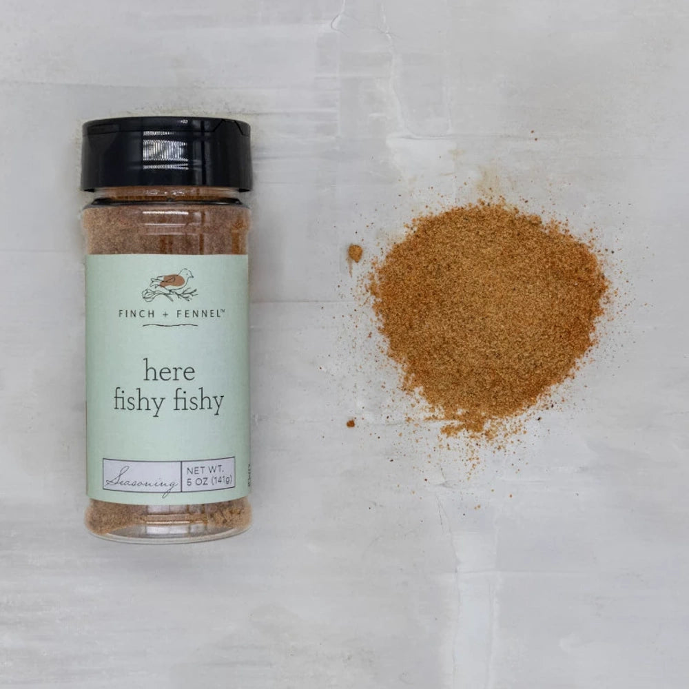 Finch + Fennel "Here Fishy Fishy" seasoning bottle next to a pile of brown spice rub on a light grey background.