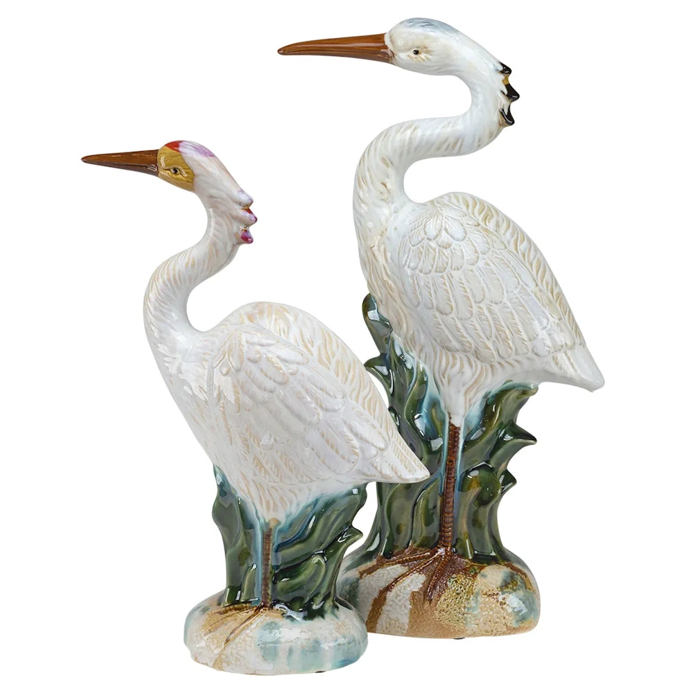 Pair of glossy ceramic heron figurines standing in green marsh grass, featuring white feathers and long beaks.