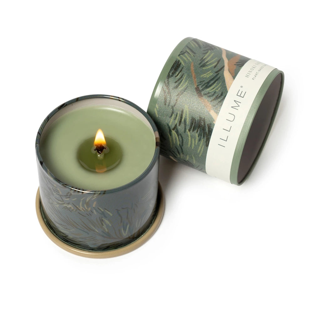 Top-down view of a lit Illume candle with green wax, placed next to its pine-patterned lid on a white backdrop.