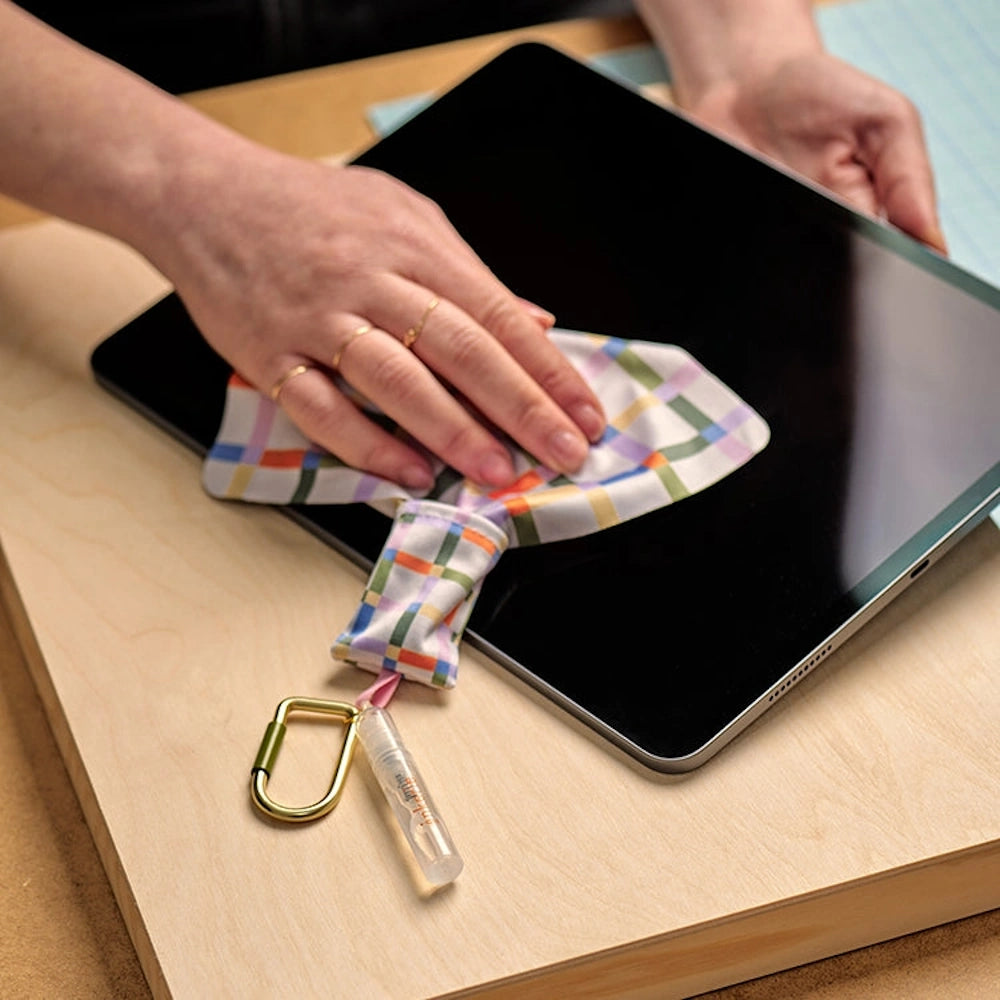Inkerie on-the-go lens and screen cleaning kit with plaid microfiber cloth and spray bottle cleaning a tablet.