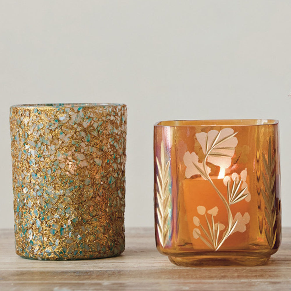 Iridescent amber glass votive holder with a lit tealight next to a gold mosaic holder on a tabletop.