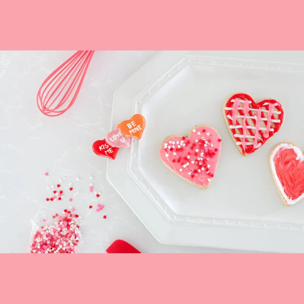 Heart-shaped sugar cookies with pink and red icing and sprinkles on a white plate with a pink whisk and candy hearts.
