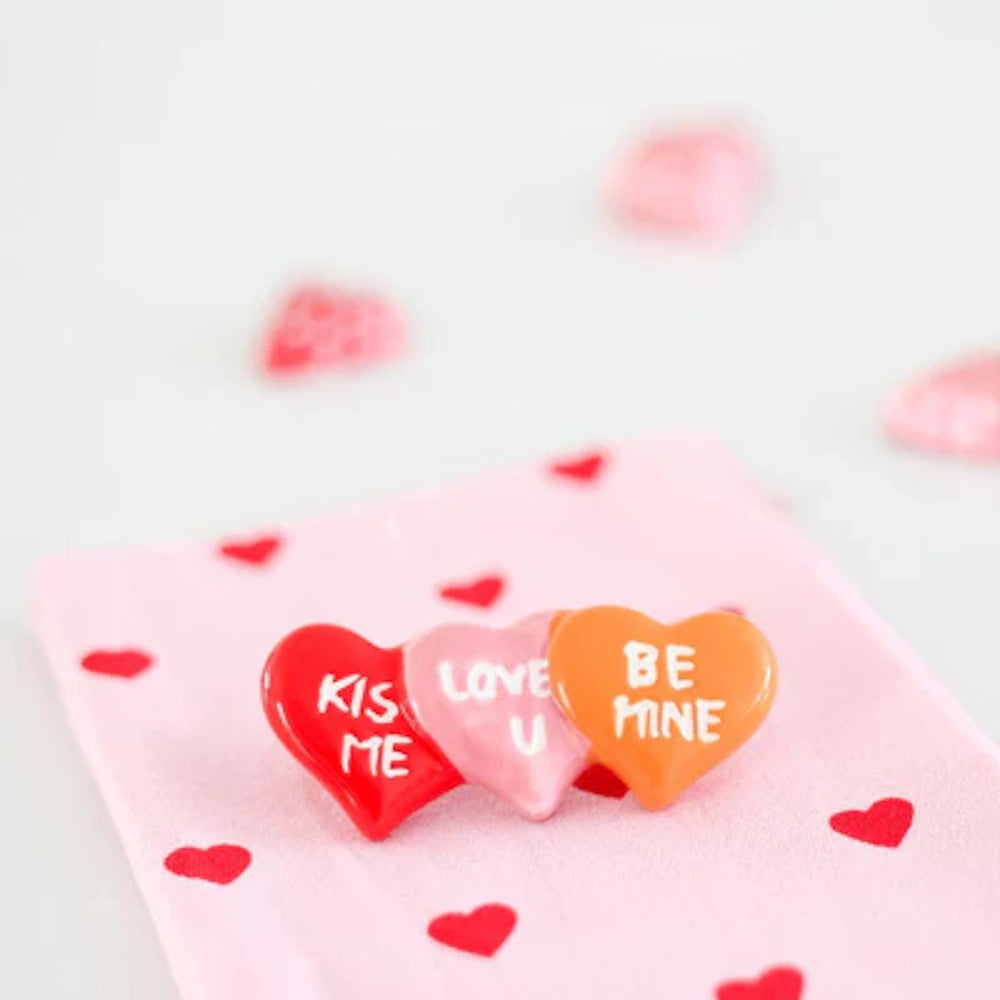Red, pink, and orange heart-shaped pins with romantic phrases resting on a pink heart-patterned fabric.
