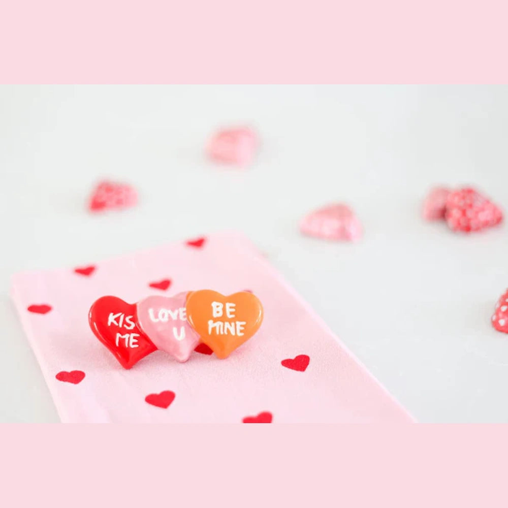 Three conversation heart chocolates on a pink cloth with the phrases Kiss Me, Love U, and Be Mine.