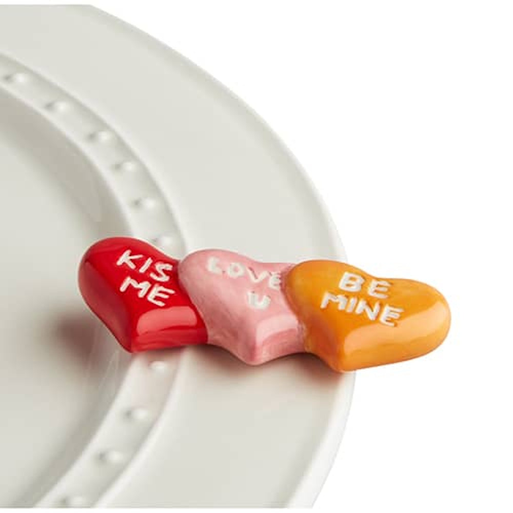 Three conversation hearts in red, pink, and orange with "Kiss Me," "Love U," and "Be Mine" on a white ceramic plate.
