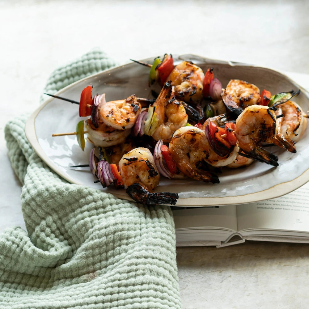 Grilled shrimp skewers with red onion and peppers served on a oval plate next to a mint green towel.