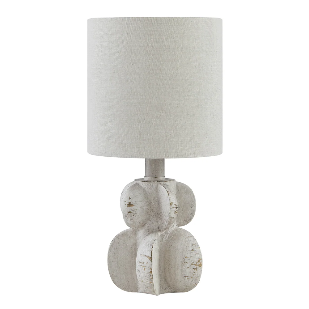 A table lamp with a white textured, sculptural base featuring organic, disk-like shapes and a beige linen drum shade.