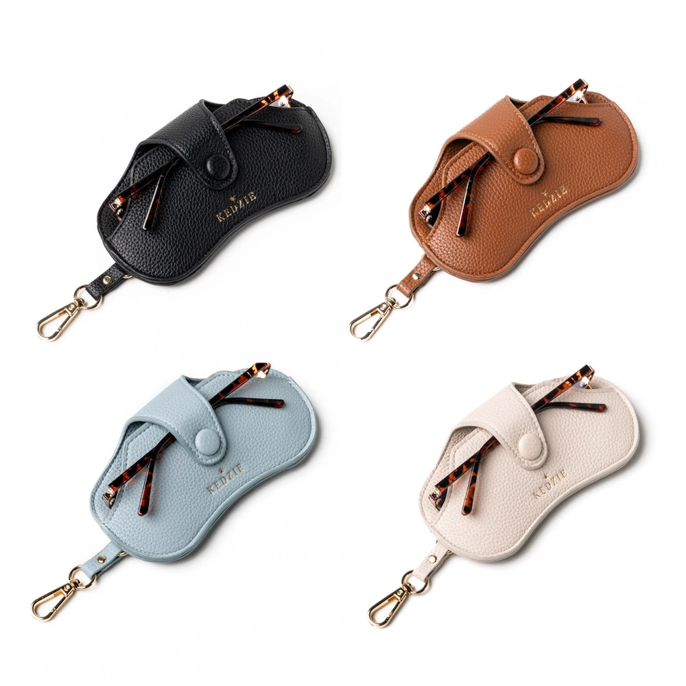 Four Kedzie pebbled leather eyewear cases in black, brown, blue, and cream with gold clips and tortoise shell glasses.