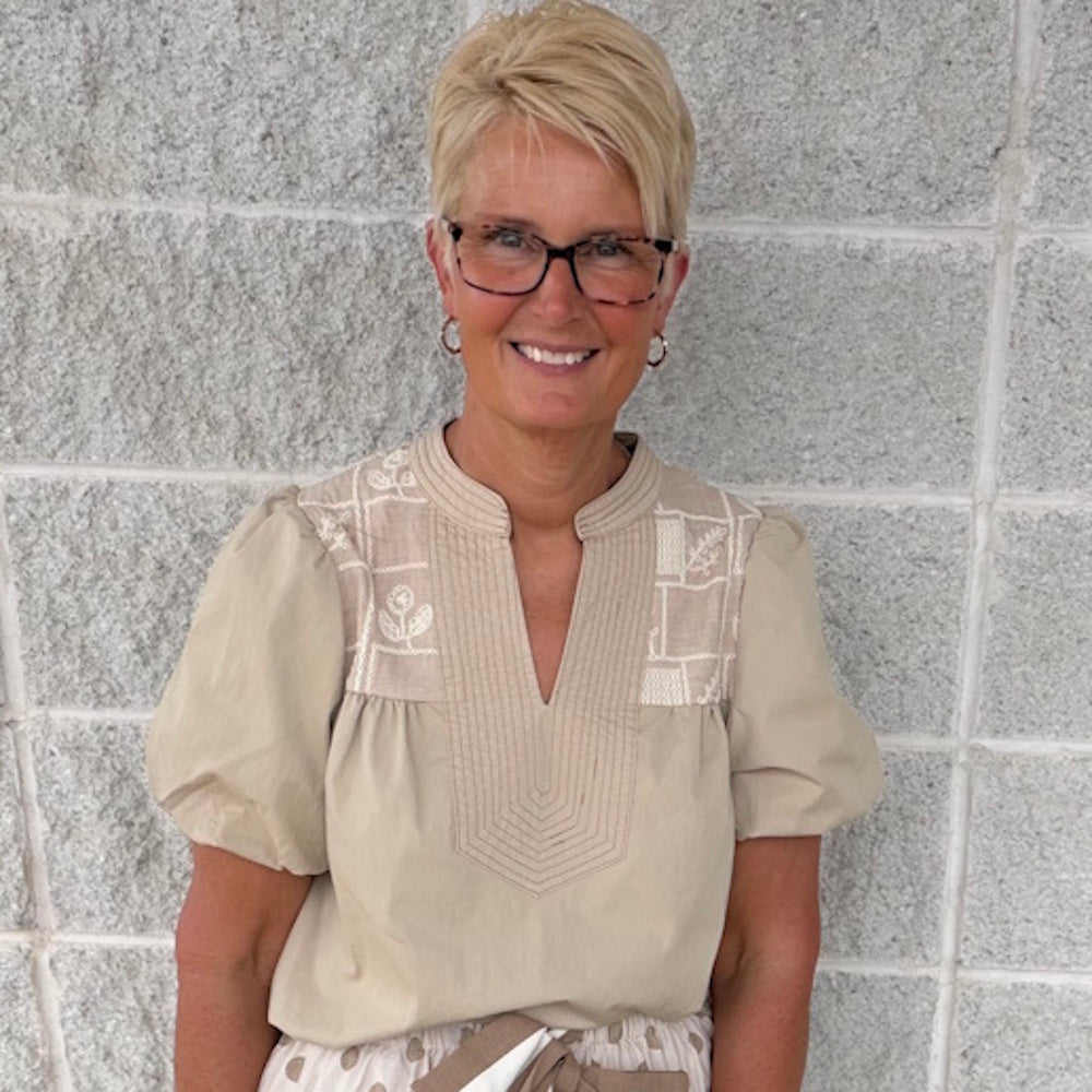 Woman with short blonde hair and glasses wearing a beige puff-sleeve top with embroidered details.