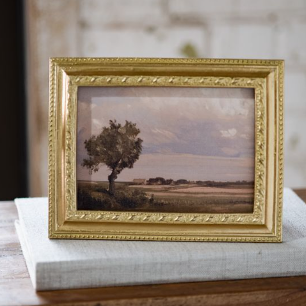 Framed landscape painting with a tree in an ornate gold frame sitting on a light gray book.