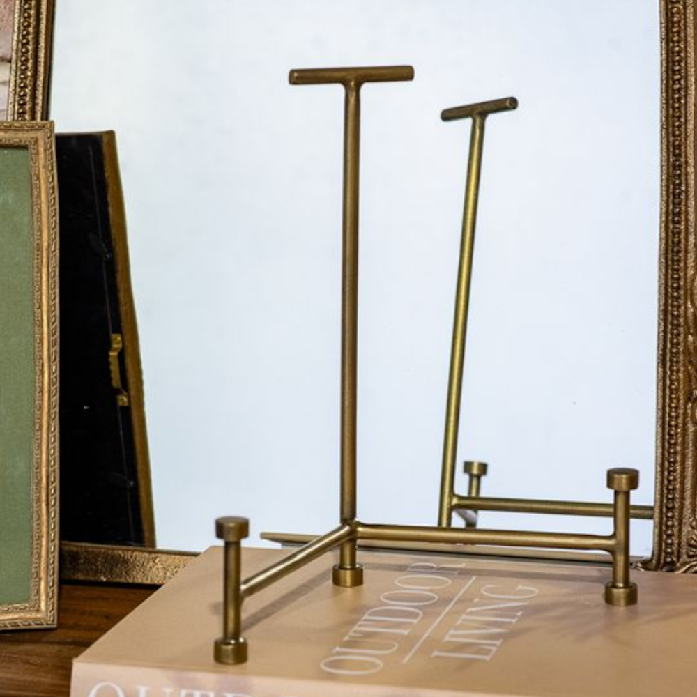 Tall antique brass display easel with a T-bar top and round-footed legs on a coffee table book.