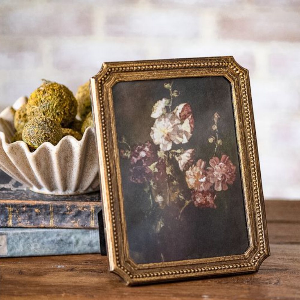 Floral still life painting in a beaded antique gold frame, set on a wooden table next to books.